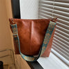 A brown leather shoulder bag with a wide teal and gold embroidered strap is displayed next to a window blind.
