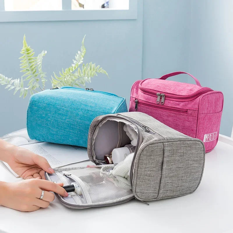 A hand opens a gray portable toiletry bag next to a pink zippered pouch on a white surface.