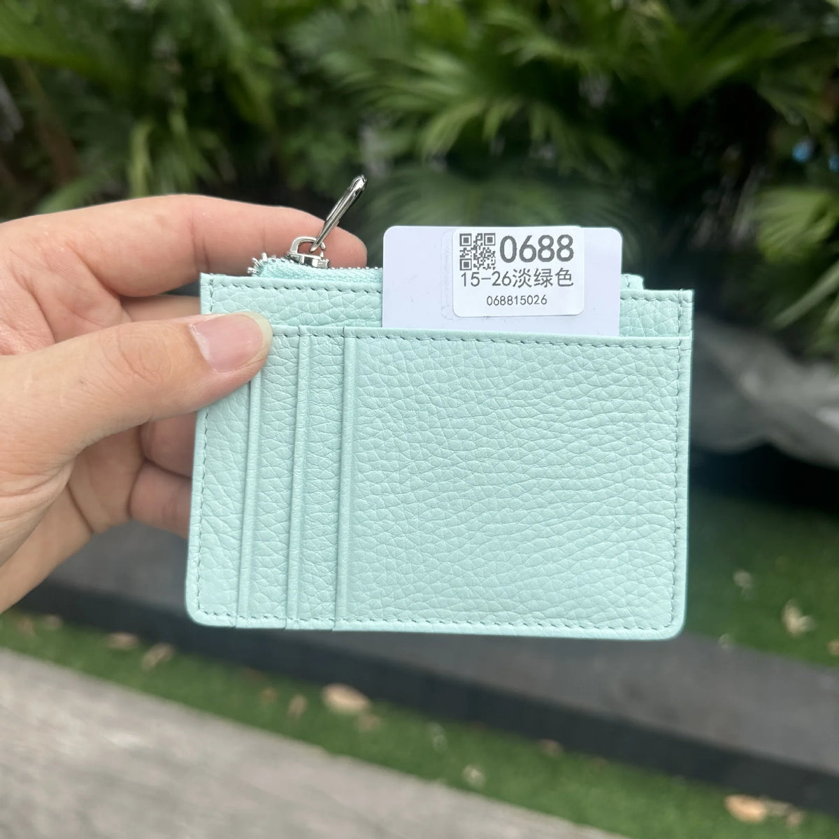 A hand holds a light mint green genuine leather zipper card holder with a silver clasp and a white tag.
