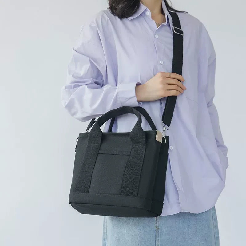 A woman is holding a black canvas crossbody bag with an adjustable strap and multiple pockets against a light purple shirt.