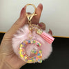 A gold-colored keychain featuring a pink faux fur pompom and a resin initial letter charm.