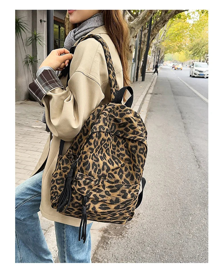 A woman wearing a beige coat and jeans carries a leopard print canvas backpack while walking on a city sidewalk.