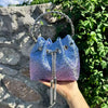 A hand holds a blue iridescent crystal-embellished evening clutch with a silver chain against a stone and green foliage backg