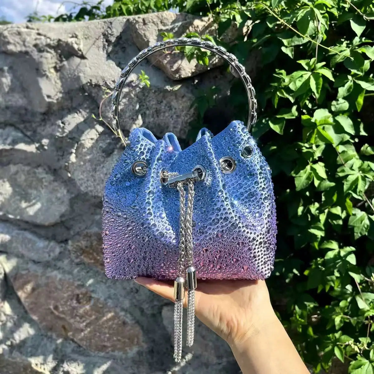 A hand holds a blue iridescent crystal-embellished evening clutch with a silver chain against a stone and green foliage backg