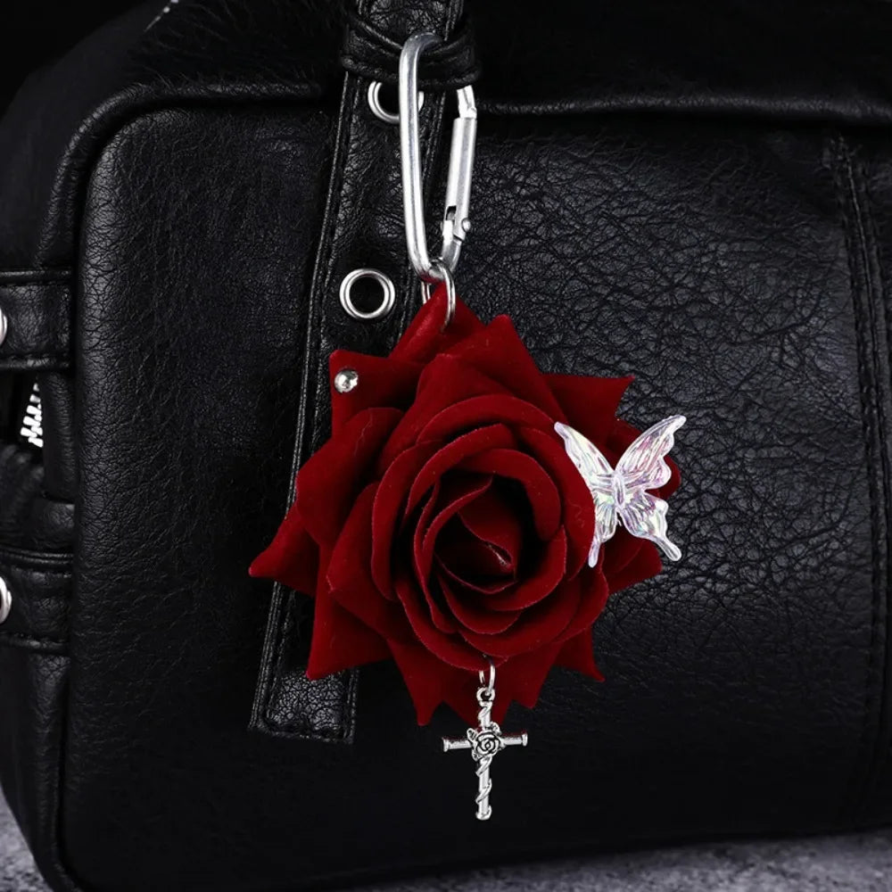 A realistic red rose flower keychain with a silver cross and a white butterfly attached to a black leather bag.
