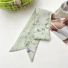 Un foulard de cheveux floral en soie vert clair plié en diagonale sur une surface blanche à côté d'une bouteille en verre.