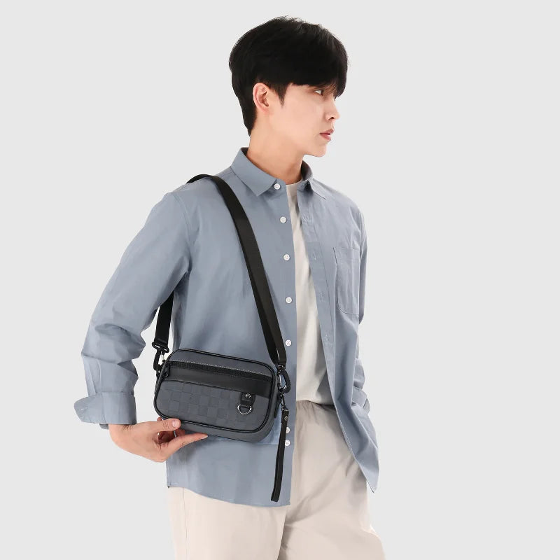 A young man wearing a light blue shirt and beige pants holds a black PU leather crossbody bag with an adjustable strap.