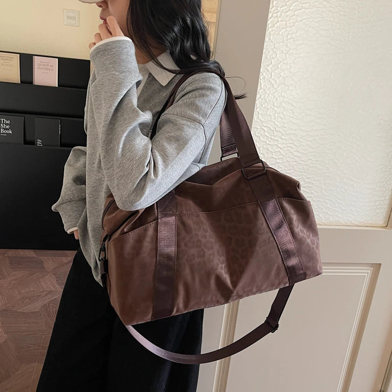 A woman holds a brown leopard print duffle bag with a leather strap against a light-colored wall.