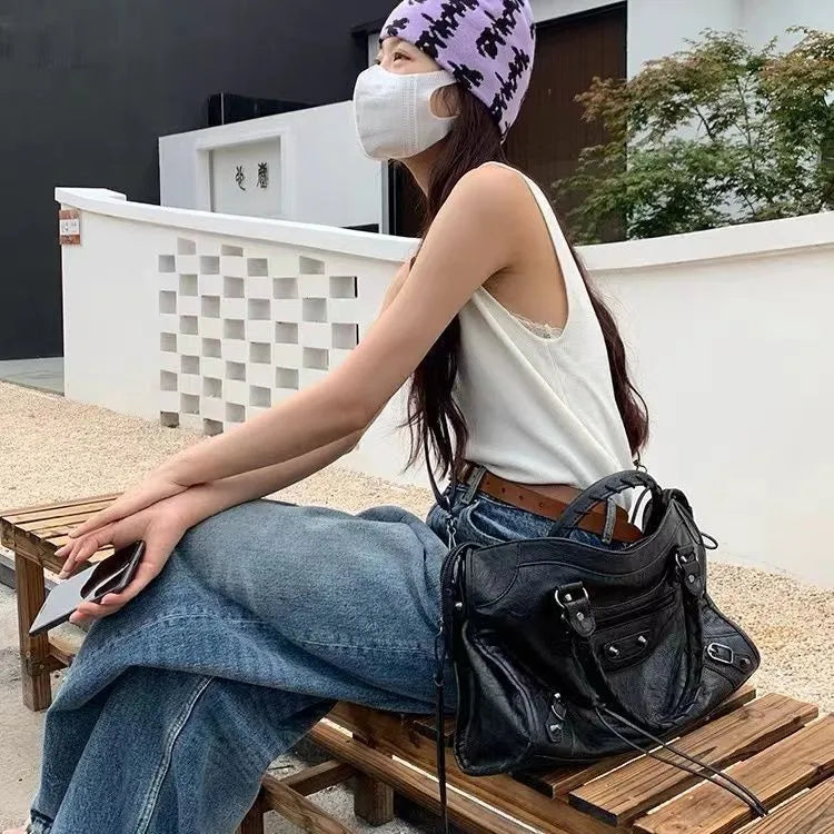 A woman wearing a white tank top and jeans sits on a wooden bench holding a black saddle-shaped PU leather city tote bag.