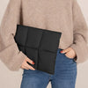 Une personne tient une housse de laptop noire en duvet contre un pull beige et un jean bleu.