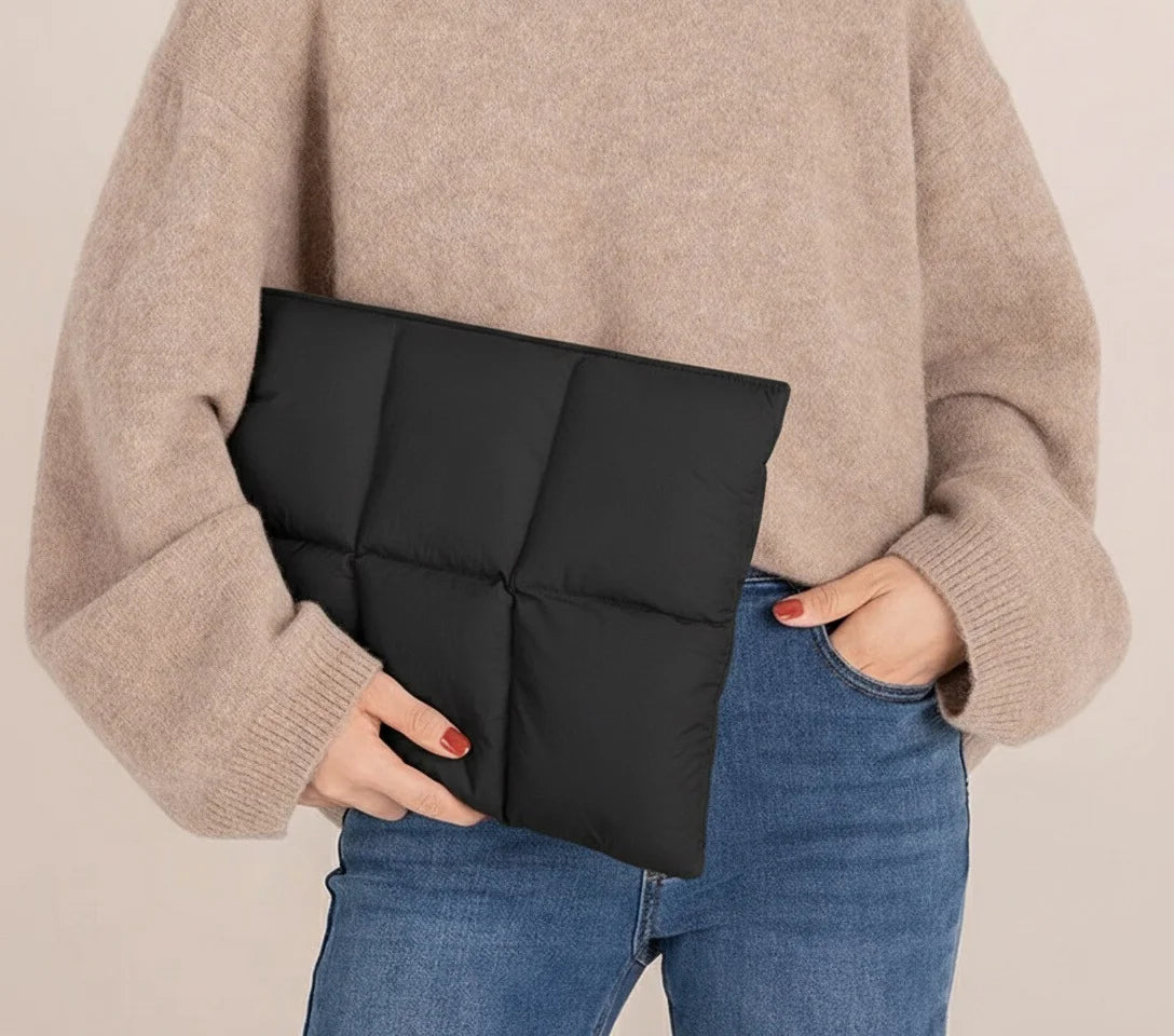 Une personne tient une housse de laptop noire en duvet contre un pull beige et un jean bleu.