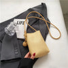 Bolso cruz de cuero beige claro con correa color canela, mostrado contra un blazer gris y un objeto de vidrio transparente.