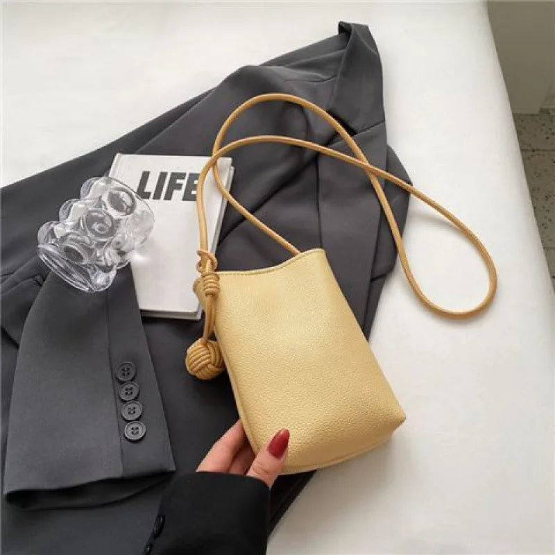 Bolso cruz de cuero beige claro con correa color canela, mostrado contra un blazer gris y un objeto de vidrio transparente.