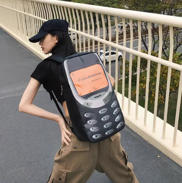 A woman wearing a black shirt and tan pants carries a black vintage Y2K telephone backpack outdoors.