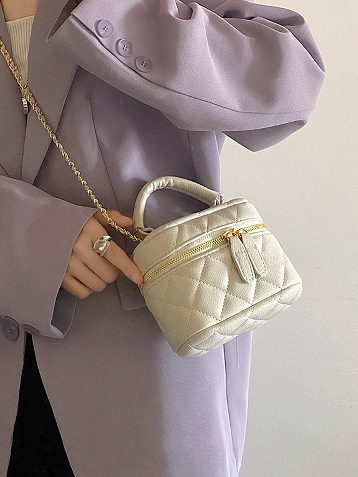 Une main tient un petit sac bandoulière matelassé beige clair avec une chaîne dorée, contre un blazer lavande.