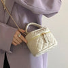 Une main tient un mini sac bandoulière beige clair matelassé avec une chaîne dorée contre un blazer lavande.