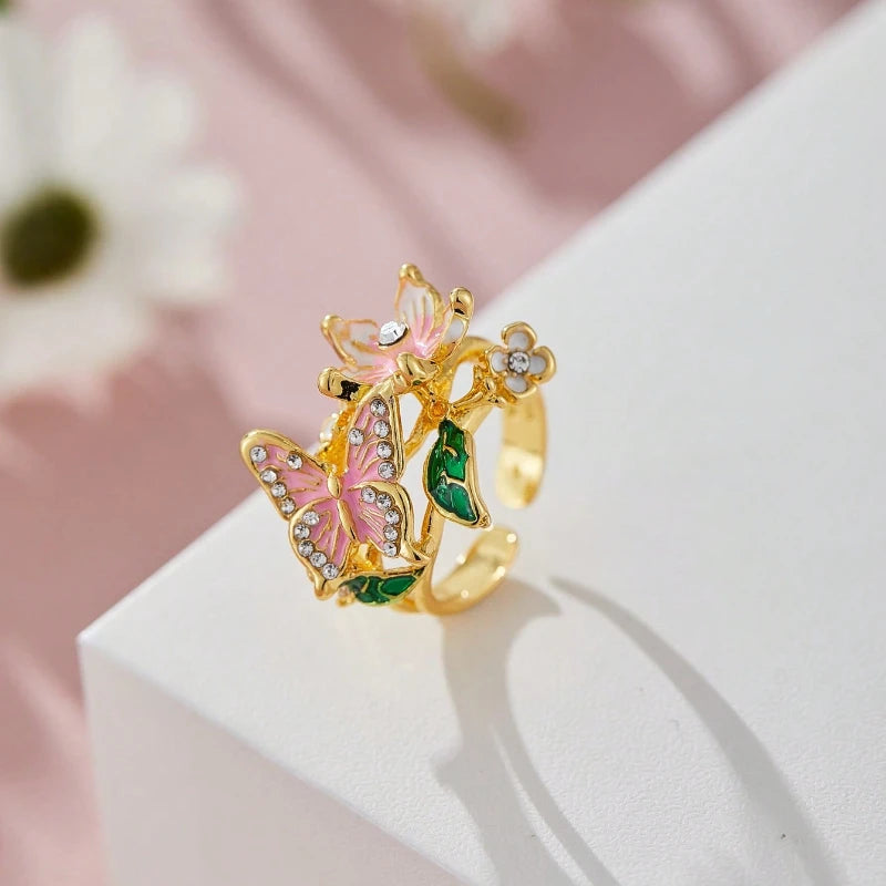 Gold-toned aesthetic flower butterfly rings with pink and green accents displayed on a white surface.