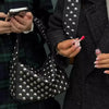 A person holds a black designer metal rivet shoulder bag and a smartphone against a dark plaid coat.