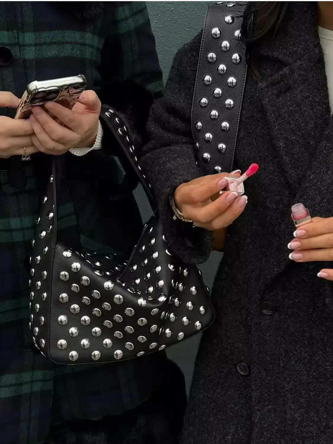 A person holds a black leather shoulder bag with silver metal rivets and a smartphone.