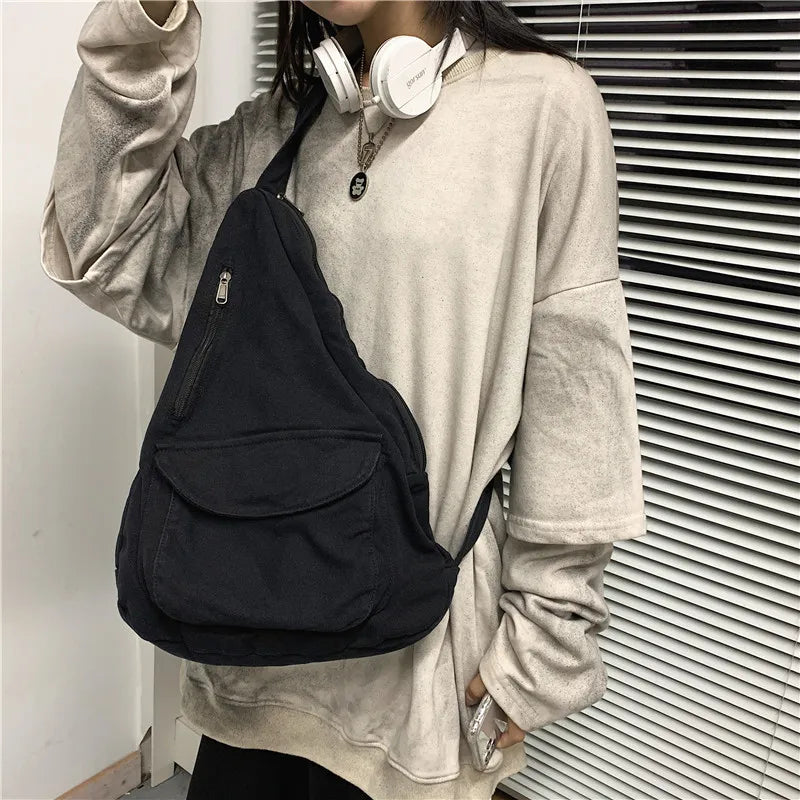 A person wearing a beige sweatshirt holds a black canvas crossbody shoulder bag with multiple pockets.