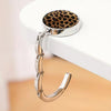 A silver zinc alloy folding handbag hook with a leopard print leather attachment resting on a white surface.