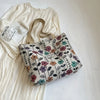 A beige bohemian corduroy tote bag with a floral pattern rests next to a white linen dress and a clear glass.