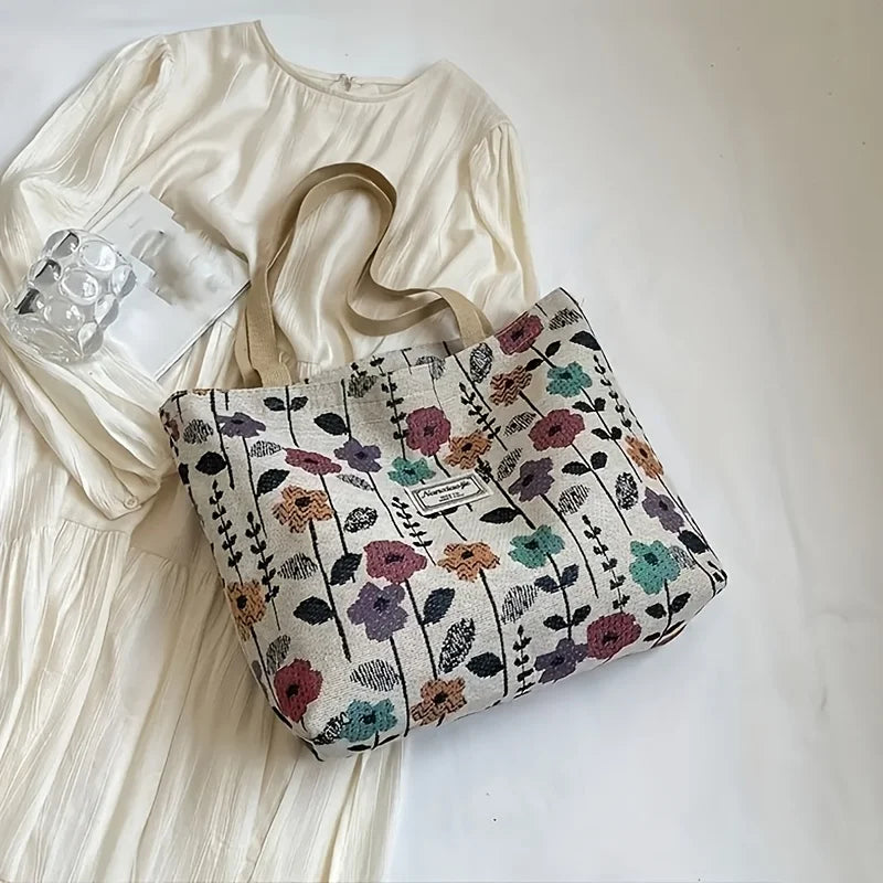 A beige bohemian corduroy tote bag with a floral pattern rests next to a white linen dress and a clear glass.