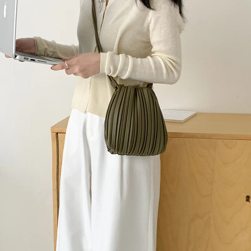 A woman wearing a cream sweater and white skirt holds a olive green pleated PU leather crossbody bag while using a laptop.