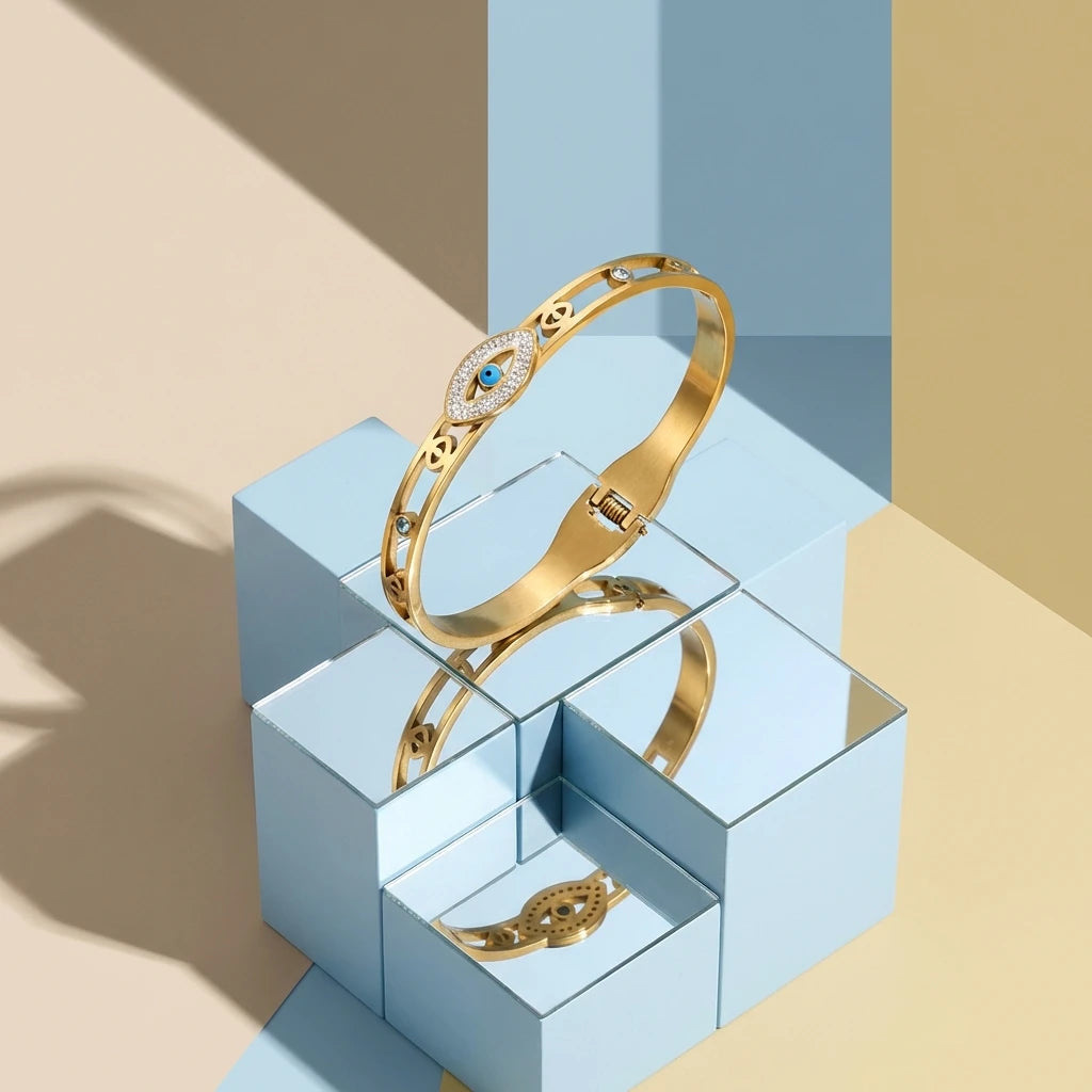 A gold cuff bracelet featuring an evil eye crystal design displayed on a geometric light blue box.