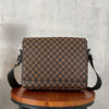 A brown patterned flap crossbody bag resting on a wooden stool against a gray background.