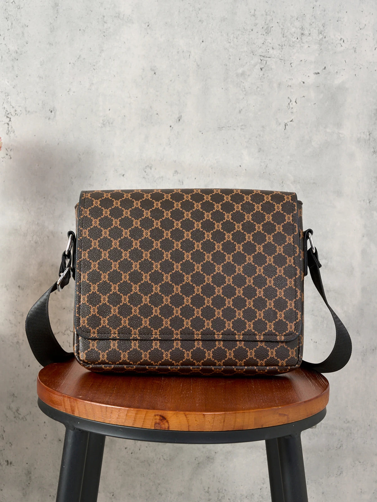 A brown patterned flap crossbody bag resting on a wooden stool against a gray background.