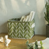 A green storage basket with a herringbone pattern holds stacked white books on a wooden surface.