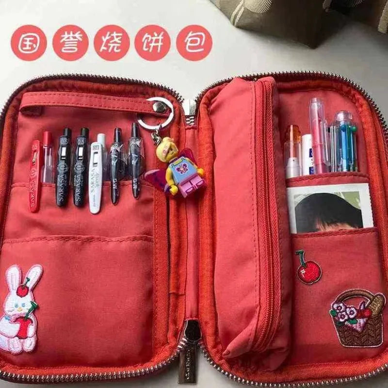 A red corduroy pencil case with a fold-out tray is shown open, displaying various stationery items inside.