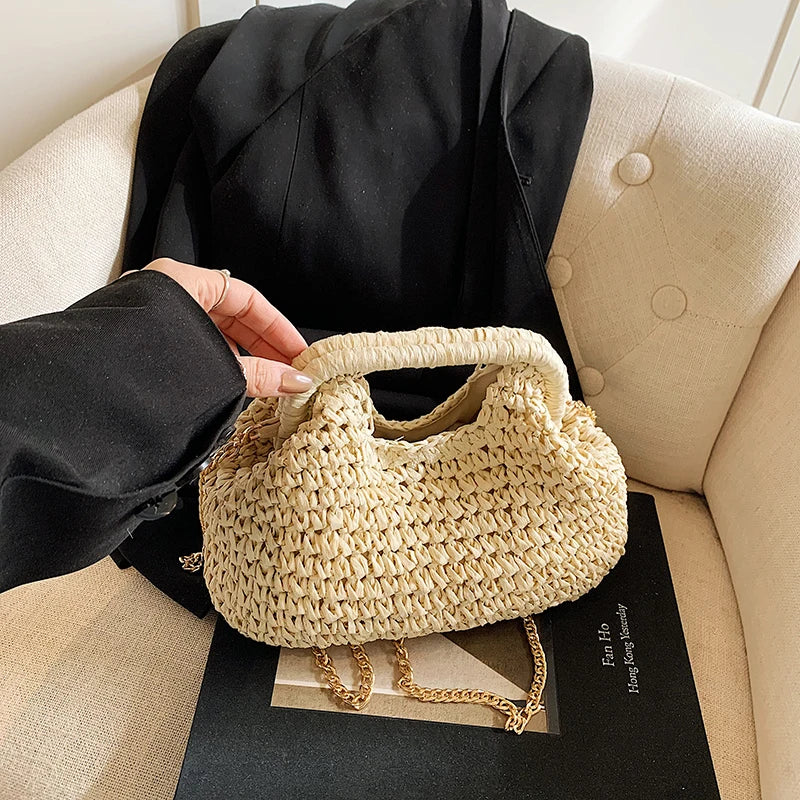 A light beige chain woven straw shoulder bag resting on a black surface next to a person's hand.
