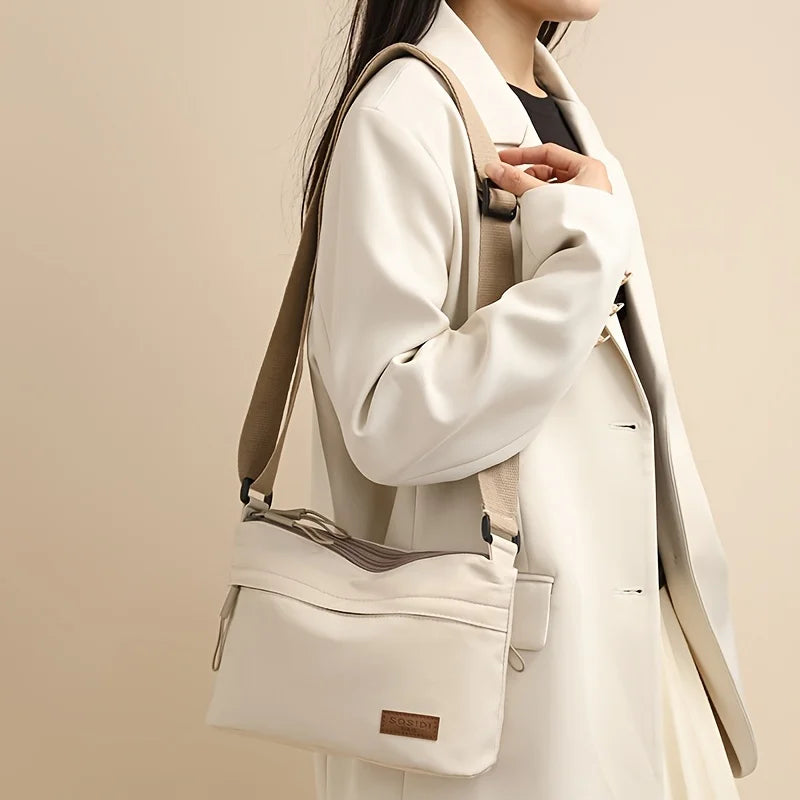 A woman holds a beige nylon crossbody shoulder bag with multiple zippered compartments against a light background.