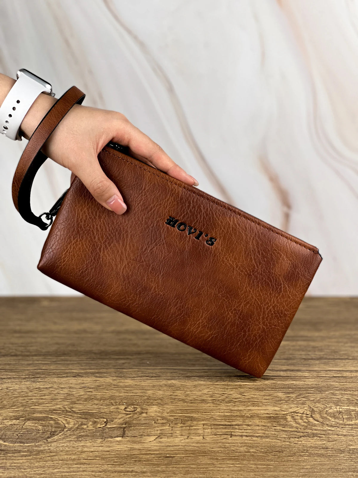 Une main tient un porte-monnaie/sac pochette en cuir marron sur une surface en bois.