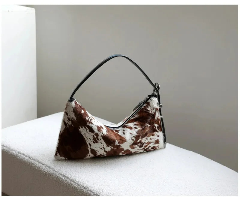 A brown and white cowhide crossbody bag with black leather strap resting on a white surface.