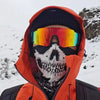 A person wearing an orange hooded jacket and a skull face mask with mirrored sunglasses in a snowy outdoor setting.
