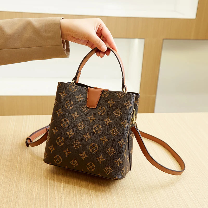 A hand holds a brown Western floral bucket bag with an adjustable leather crossbody strap on a light wooden surface.