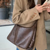 A woman holds a brown PU leather tote bag with a crossbody strap under a beige blazer.