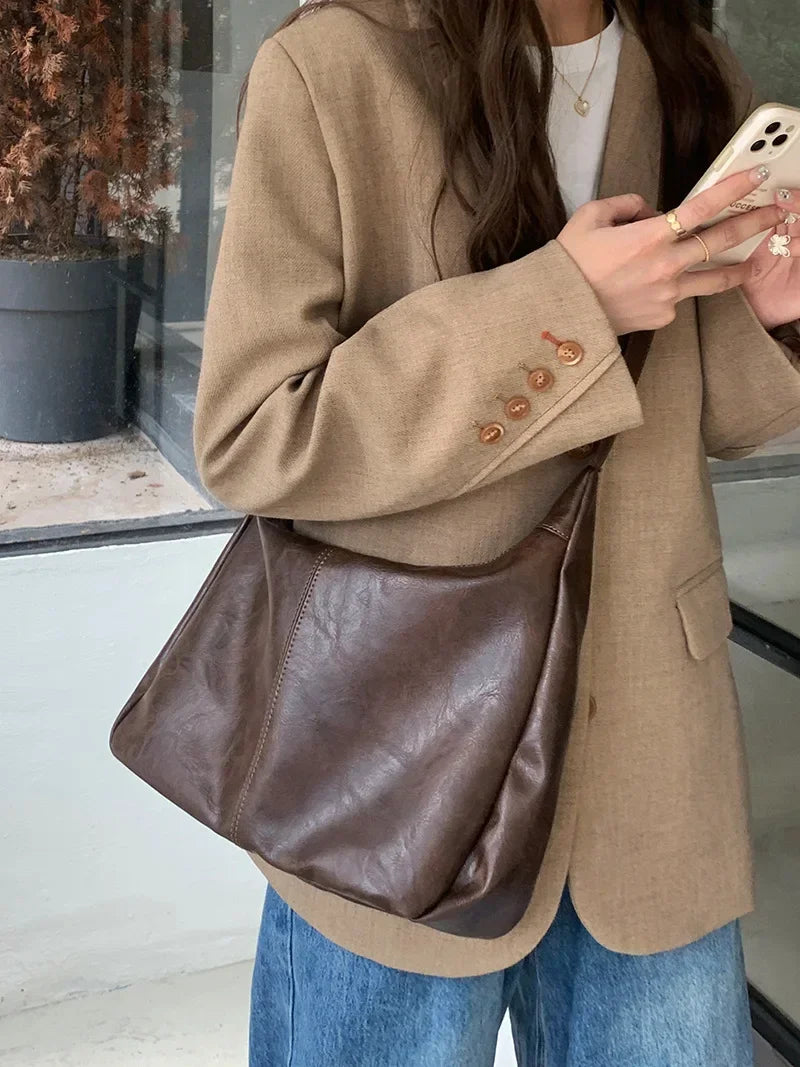 A woman holds a brown PU leather tote bag with a crossbody strap under a beige blazer.