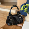 A black PU leather shoulder bag with silver rivets resting on a wooden surface next to a white vase of green flowers.