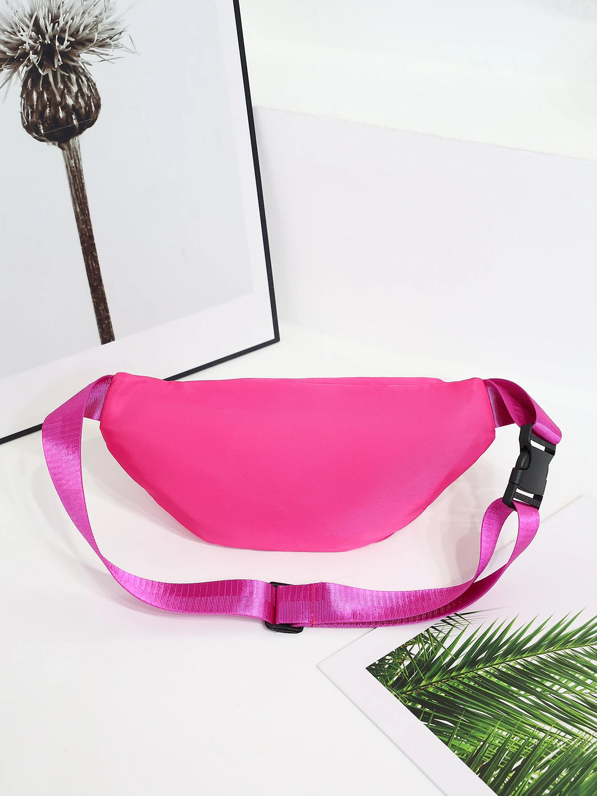 A bright pink multi-layer sports waist bag is displayed on a white surface next to a framed feather and a green palm leaf.