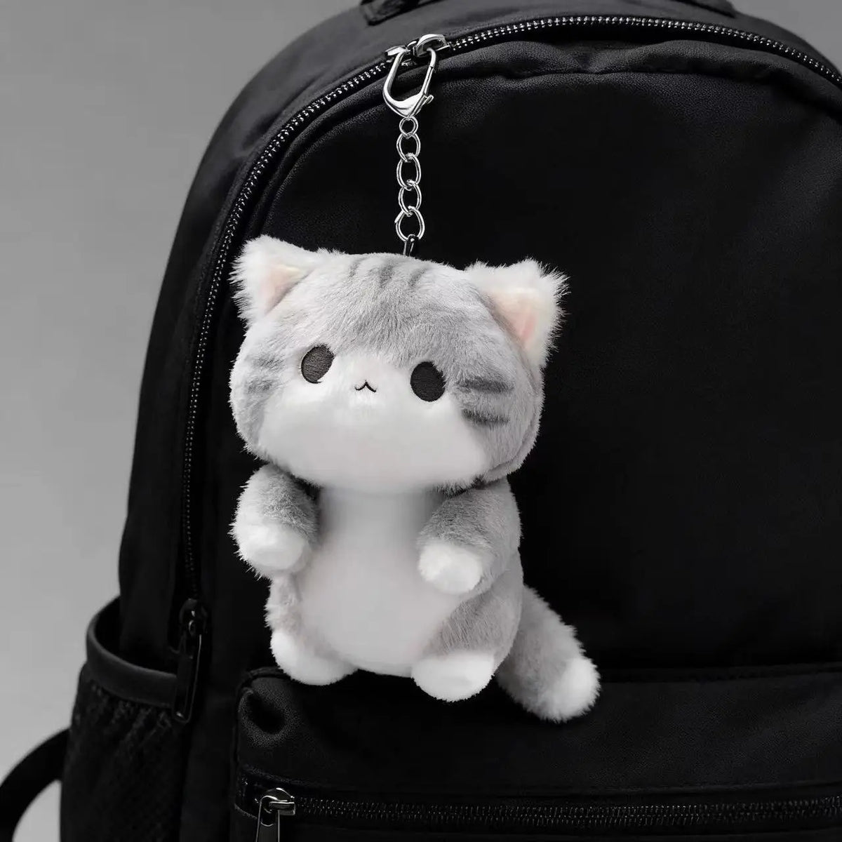 A grey soft cat plush pendant ornament attached to a black backpack.