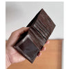 A hand holds a dark brown genuine leather bifold wallet displaying stitched lines and a suede flap.