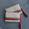 Beige und grün gemusterte Clutch-Tasche mit schwarzem Zierrand auf blauem Stoffhintergrund.