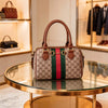 A brown leather tote diaper bag with a Gucci monogram pattern and green and red stripes sits on a glass table.