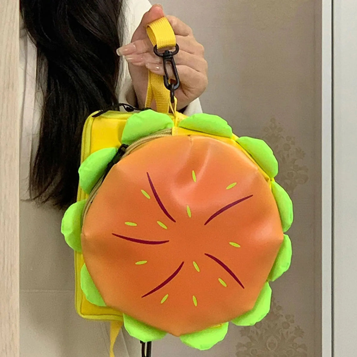 A bright orange and green novelty backpack shaped like a sliced tomato, being held by a person's hand.