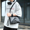 A man wearing a gray t-shirt and black accessories holds a black embossed crossbody clutch bag featuring a pistol design.