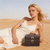A blonde woman lies in a desert landscape next to a gray double-layer makeup storage case.
